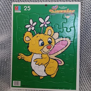 Könnte beinhalten: Ein grünes Puzzlespiel mit einem Cartoon-Bären mit Schmetterlingsflügeln und Blumenakzenten. Das Puzzle ist mit "MB 25" und "Walt Disney Presents Tuzzlers" beschriftet. Empfohlen für Kinder im Alter von 3 bis 7 Jahren.