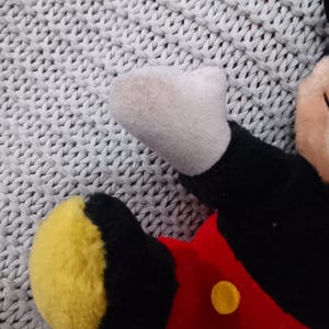 K&ouml;nnte beinhalten: Ein Pl&uuml;sch-Mickey-Mouse-Spielzeug mit schwarzem Fell, roten Shorts mit gelben Kn&ouml;pfen und gelben Schuhen. Das Spielzeug liegt auf einer grauen gestrickten Oberfl&auml;che.