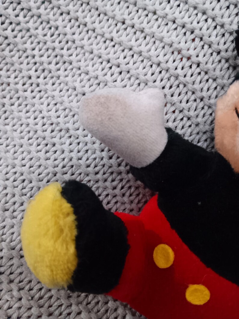 K&ouml;nnte beinhalten: Ein Pl&uuml;sch-Mickey-Mouse-Spielzeug mit schwarzem Fell, roten Shorts mit gelben Kn&ouml;pfen und gelben Schuhen. Das Spielzeug liegt auf einer grauen gestrickten Oberfl&auml;che.