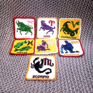 May include: Seven embroidered zodiac patches. Each patch is square with a white background and a coloured border. The signs include Taurus, Aries, Capricorn, Pisces, Virgo, Aquarius, and Scorpio. The patches are displayed on a grey knitted surface.