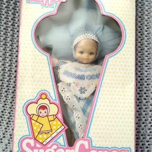 May include: A vintage box for a Baby Moppets Sugar Cones doll. The box is pink, yellow, and blue with a white background. The doll is wearing a blue and white gingham dress and a white bonnet. The box has the words "Baby Moppets" and "Sugar Cones" on it.