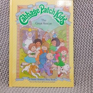 Könnte beinhalten: Ein gelber Buchumschlag mit dem Titel "Cabbage Patch Kids The Great Rescue" und einem Bild einer Gruppe von Cabbage Patch Kids Puppen mit ihren Haustieren. Das Buch ist ein Parker Brothers Story Book mit einer Geschichte von Mark Taylor und Bildern von Jan Brett.
