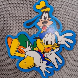 May include: A blue, yellow, and white cartoon cutout of Donald Duck and Goofy. Donald Duck is wearing a blue hat and Goofy is wearing a yellow hat. They are both smiling and have their arms outstretched.