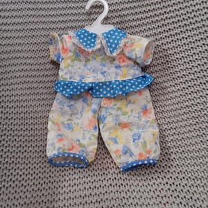 Könnte beinhalten: Puppenkleidung-Set mit einem Oberteil und einer Hose mit floralem Muster, mit blauen Tupfen verziert. Das Oberteil hat einen weißen Spitzenkragen und blaue Tupfen-Details. Das Outfit befindet sich auf einem weißen Kleiderbügel.