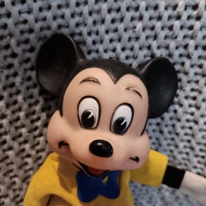May include: A vintage Mickey Mouse figurine. The figure has large black ears, a black nose, and a smiling expression. Mickey wears a yellow jacket, red pants, a blue bow tie, and black gloves. A red button is visible on the jacket.