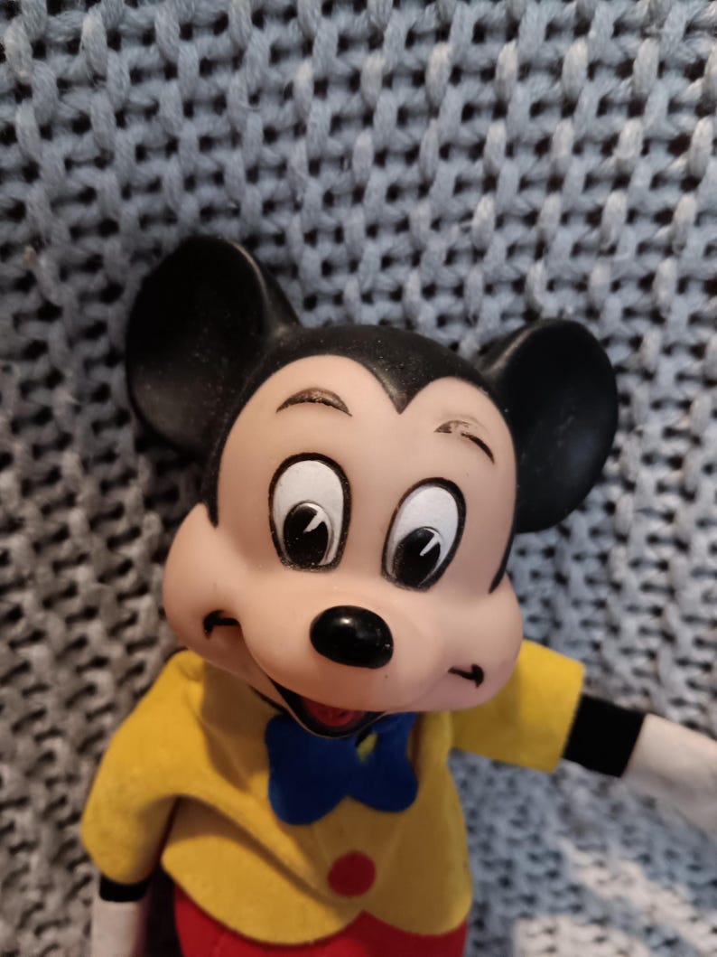May include: A vintage Mickey Mouse figurine. The figure has large black ears, a black nose, and a smiling expression. Mickey wears a yellow jacket, red pants, a blue bow tie, and black gloves. A red button is visible on the jacket.