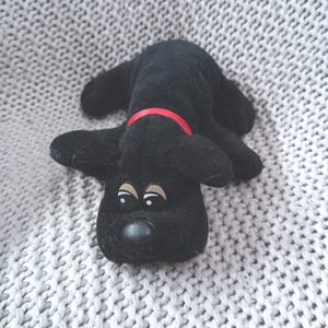 Könnte beinhalten: Ein schwarzes Plüsch-Welpenspielzeug mit rotem Halsband. Das Spielzeug hat große, ausdrucksstarke Augen und eine schwarze Nase. Der Welpe liegt auf einer grauen, strukturierten Oberfläche.