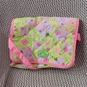 May include: A Cabbage Patch Kids shoulder bag with a light green quilted background. The bag features pink and lavender flowers, a pink strap, and pink trim. The bag has the words "Cabbage Patch Kids" printed on it.