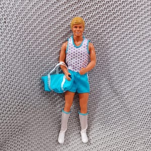Puede incluir: Una muñeca vintage con cabello rubio que lleva un top blanco sin mangas con un patrón de estrellas, pantalones cortos azules y botas blancas. La muñeca sostiene una bolsa de mano azul y blanca.