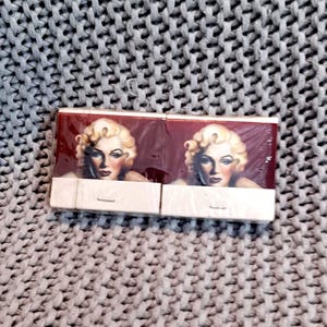 May include: Two matchbooks featuring a portrait of Marilyn Monroe. The matchbooks have a burgundy background and a white striker strip. The image is set against a gray knitted background.