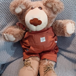 Puede incluir: Un oso de peluche marrón con un mono color óxido y botas de trabajo color beige. El oso tiene una cara beige claro y una nariz marrón oscuro. El mono tiene un pequeño bolsillo con un logotipo. El oso posa con los brazos extendidos.