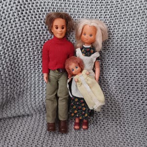 May include: A vintage doll family, including a man in a red sweater and green pants, a woman in a floral dress, and a child. The dolls have molded plastic faces and rooted hair. The family is posed together on a gray knitted surface.