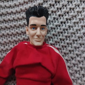 May include: A red and white toy figure of a man with black hair. The figure is wearing a red sweater with white stripes on the sleeves. The figure has a smiling face with blue eyes.