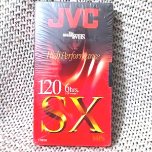 May include: A JVC VHS video cassette tape with a red and black gradient design. The tape has the text "High Performance" and "120 6hrs. SX" in gold lettering. The JVC logo is at the top.