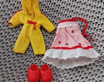 May include: A set of doll clothes including a yellow hooded jumpsuit, a pink and white dress with strawberries, and a pair of red shoes.