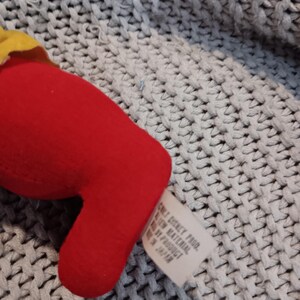 May include: A red plush toy with a yellow accent. A white tag with text is attached. The toy is on a gray knitted surface. The tag reads "ONLY DISNEY PROD. ALL NEW MATERIAL MADE BY PRODUCT IN JAPAN."