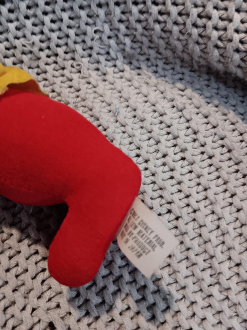 May include: A red plush toy with a yellow accent. A white tag with text is attached. The toy is on a gray knitted surface. The tag reads "ONLY DISNEY PROD. ALL NEW MATERIAL MADE BY PRODUCT IN JAPAN."