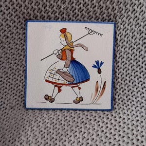 Könnte beinhalten: Quadratische Keramikfliese mit einer handgemalten Illustration einer Person in traditioneller Kleidung, die eine Harke trägt. Die Figur trägt einen blauen Rock, eine braune Jacke und einen roten Hut. Die Fliese hat einen blauen Rand.