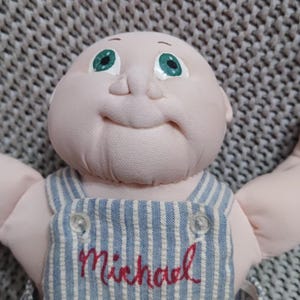 Puede incluir: Una muñeca hecha a mano con una cara suave y rosada y ojos verdes. La muñeca lleva un peto a rayas azules y blancas con el nombre "Michael" bordado en rojo. Los brazos de la muñeca están extendidos y tiene una expresión sonriente.