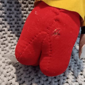 May include: Close-up of a red plush toy foot with a visible hole and stitched seams. The foot is part of a larger toy, with a yellow fabric visible above. The toy rests on a gray and white textured surface.