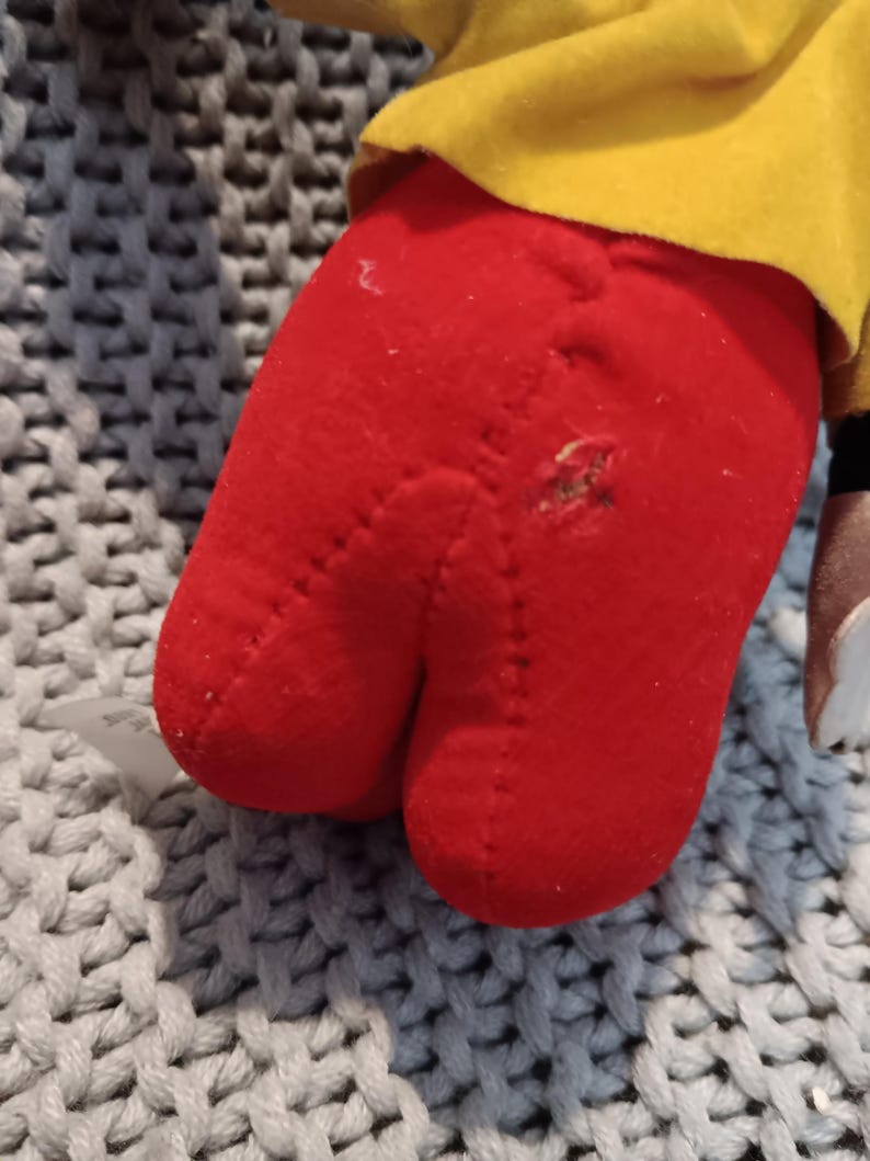 May include: Close-up of a red plush toy foot with a visible hole and stitched seams. The foot is part of a larger toy, with a yellow fabric visible above. The toy rests on a gray and white textured surface.