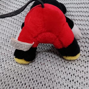 K&ouml;nnte beinhalten: Ein rotes und schwarzes Pl&uuml;schtier von Mickey Mouse. Das Spielzeug liegt auf dem R&uuml;cken mit den Beinen in der Luft. Das Spielzeug hat einen schwarzen Schwanz und gelbe Schuhe.