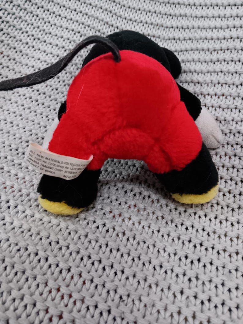 K&ouml;nnte beinhalten: Ein rotes und schwarzes Pl&uuml;schtier von Mickey Mouse. Das Spielzeug liegt auf dem R&uuml;cken mit den Beinen in der Luft. Das Spielzeug hat einen schwarzen Schwanz und gelbe Schuhe.
