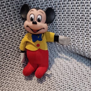 May include: A Mickey Mouse plush toy. The toy has a black head with large ears, a yellow jacket, red pants, and white gloves. A blue bow tie and a red button are also visible. The plush is sitting on a gray knitted surface.