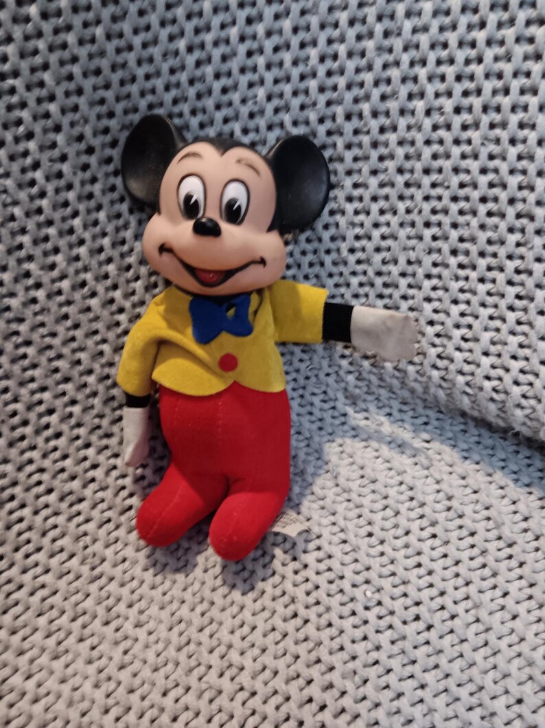May include: A Mickey Mouse plush toy. The toy has a black head with large ears, a yellow jacket, red pants, and white gloves. A blue bow tie and a red button are also visible. The plush is sitting on a gray knitted surface.