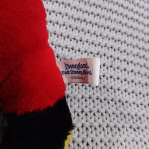K&ouml;nnte beinhalten: Rotes Pl&uuml;schtier mit schwarzen und gelben Akzenten. Das Spielzeug hat ein Etikett mit der Aufschrift "Disneyland Walt Disney World &copy;DISNEY".
