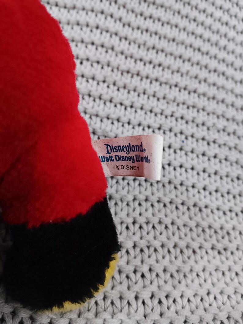 K&ouml;nnte beinhalten: Rotes Pl&uuml;schtier mit schwarzen und gelben Akzenten. Das Spielzeug hat ein Etikett mit der Aufschrift "Disneyland Walt Disney World &copy;DISNEY".