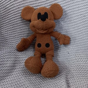 K&ouml;nnte beinhalten: Ein braunes Pl&uuml;schtier einer Zeichentrickmaus mit gro&szlig;en Ohren, schwarzen Augen und einem l&auml;chelnden Gesicht. Das Spielzeug hat ein flauschiges Aussehen und liegt auf einer grauen Strickdecke.