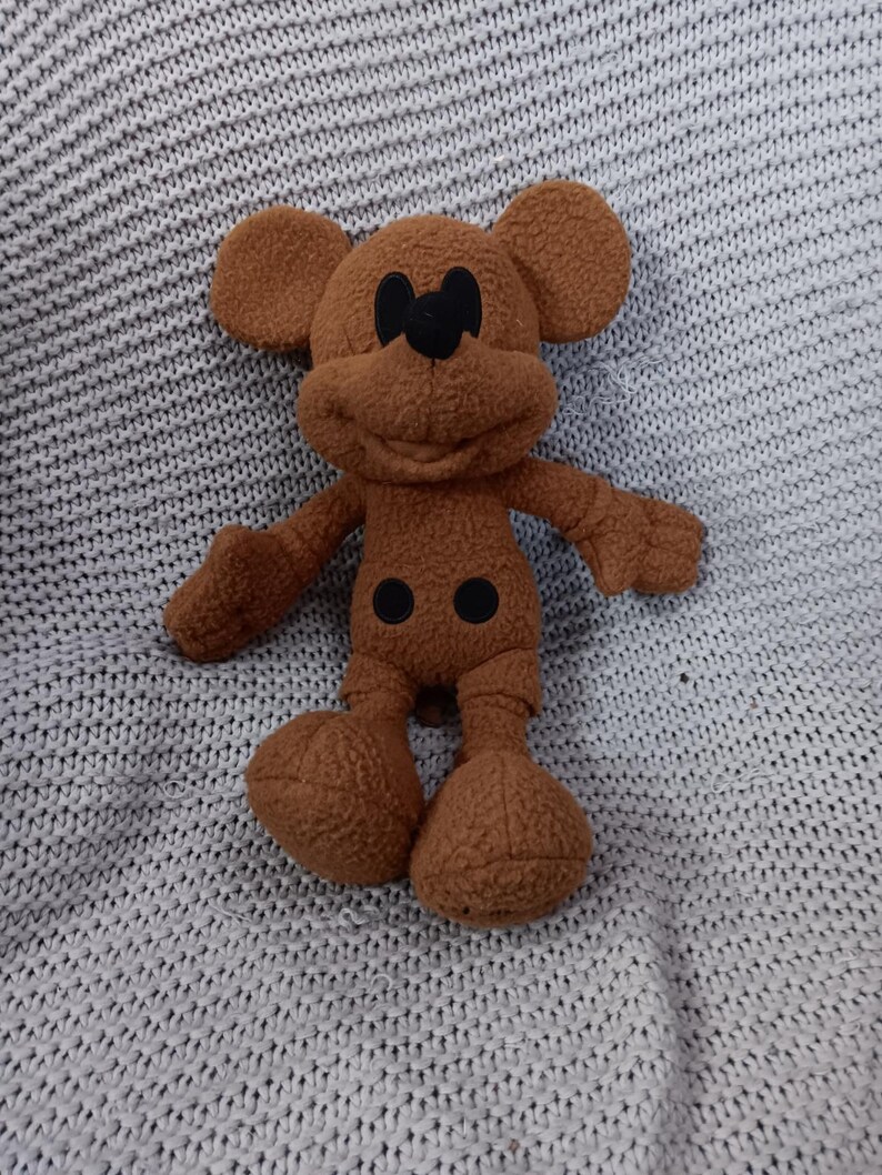 K&ouml;nnte beinhalten: Ein braunes Pl&uuml;schtier einer Zeichentrickmaus mit gro&szlig;en Ohren, schwarzen Augen und einem l&auml;chelnden Gesicht. Das Spielzeug hat ein flauschiges Aussehen und liegt auf einer grauen Strickdecke.
