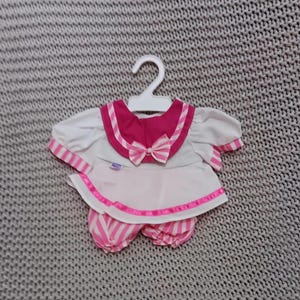 Puede incluir: Un conjunto de muñeca bebé a rayas blancas y rosas con un lazo rosa y un cuello de marinero. El conjunto está colgado en una percha blanca.