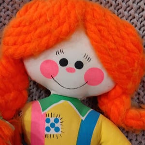 May include: A handmade doll with a white face, pink cheeks, and a smiling mouth. The doll has bright orange yarn hair styled in pigtails. It wears a yellow shirt with a blue and pink stripe, and blue overalls. A vintage toy.