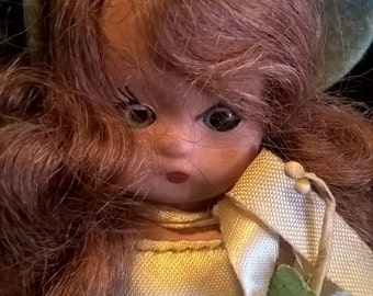 Puede incluir: Una muñeca de porcelana con cabello rosado, vestida con un vestido blanco de encaje con detalles de flores azules. La muñeca lleva un sombrero azul y tiene una expresión seria.