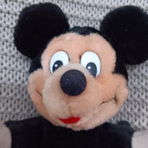 K&ouml;nnte beinhalten: Ein Pl&uuml;schtier Mickey Mouse in Schwarz und Braun mit gro&szlig;en wei&szlig;en Augen und einem roten Mund. Das Spielzeug sitzt auf einer grauen gestrickten Oberfl&auml;che.