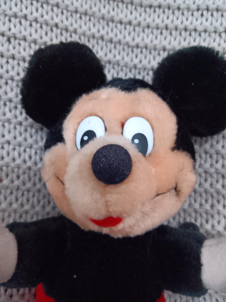 K&ouml;nnte beinhalten: Ein Pl&uuml;schtier Mickey Mouse in Schwarz und Braun mit gro&szlig;en wei&szlig;en Augen und einem roten Mund. Das Spielzeug sitzt auf einer grauen gestrickten Oberfl&auml;che.