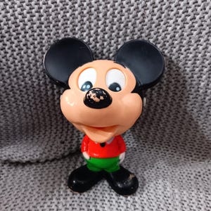 Puede incluir: Un juguete de plástico vintage de Mickey Mouse con orejas negras, una camisa roja y pantalones verdes. El juguete tiene una cara sonriente con ojos negros y una nariz negra.