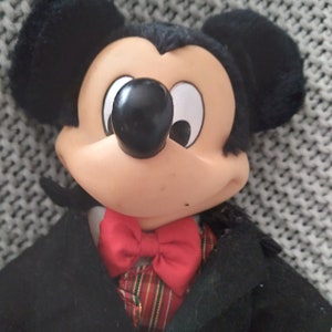 May include: A plush Mickey Mouse doll wearing a black tuxedo with a red bow tie and a green and red patterned vest.