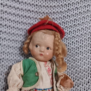 May include: A vintage doll with blonde hair, wearing a red beret, a green jacket, and a colorful patterned skirt. The doll has a painted face with blue eyes and is posed against a light blue knitted background.