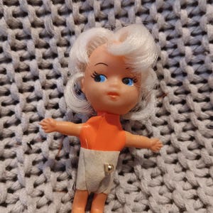 May include: A small plastic doll with white hair, blue eyes, and an orange top. The doll is wearing a diaper and has its arms outstretched. The doll is lying on a gray knitted surface.