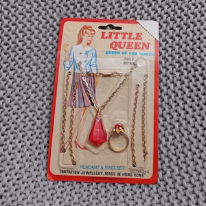 Puede incluir: Un juego de joyería de imitación para niños, que incluye un collar con un colgante rojo y un anillo rojo. El empaque presenta a una niña de dibujos animados con un vestido y el texto "Little Queen, Queen of the Month, July Ruby, No. 76529G, Pendant & Ring Set, "Imitation Jewellery. Made in Hong Kong", For 3 years and over."