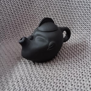 Könnte beinhalten: Eine schwarze Keramik-Teekanne mit Gesichtsdesign. Die Teekanne hat einen Ausdruck mit geschlossenen Augen, einen kleinen Ausguss und einen Griff. Der Deckel ist wie ein Hut geformt. Die Teekanne steht auf einer grauen Strickoberfläche.