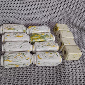 May include: A collection of ceramic drawer pulls and knobs. The drawer pulls are rectangular with white, yellow, and green marbling. The knobs are square with speckled yellow and green accents. The hardware is arranged on a gray knitted surface.