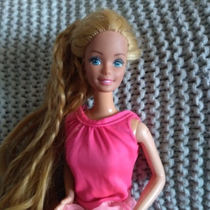May include: A vintage Barbie doll with long blonde hair, wearing a pink sleeveless top and a pink skirt. The doll has blue eyes and a painted smile.
