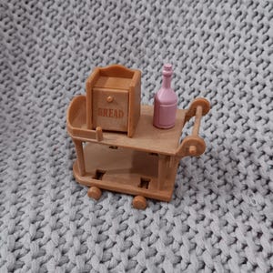 Puede incluir: Un carrito de servicio marrón en miniatura con dos estantes, que contiene una caja de pan y una botella rosa. La caja de pan tiene la palabra "BREAD" impresa. El carrito tiene ruedas pequeñas y un asa.