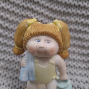 May include: A porcelain figurine of a girl with red hair in pigtails wearing a yellow swimsuit and holding a blue towel and a blue bag.