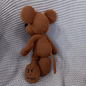 K&ouml;nnte beinhalten: Ein braunes Mickey Mouse-Pl&uuml;schtier mit gro&szlig;en, runden Ohren, einer schwarzen Nase und einem langen Schwanz. Auf einem Fu&szlig; des Spielzeugs steht "AE x Disney". Das Spielzeug liegt auf einer hellgrauen Strickoberfl&auml;che.