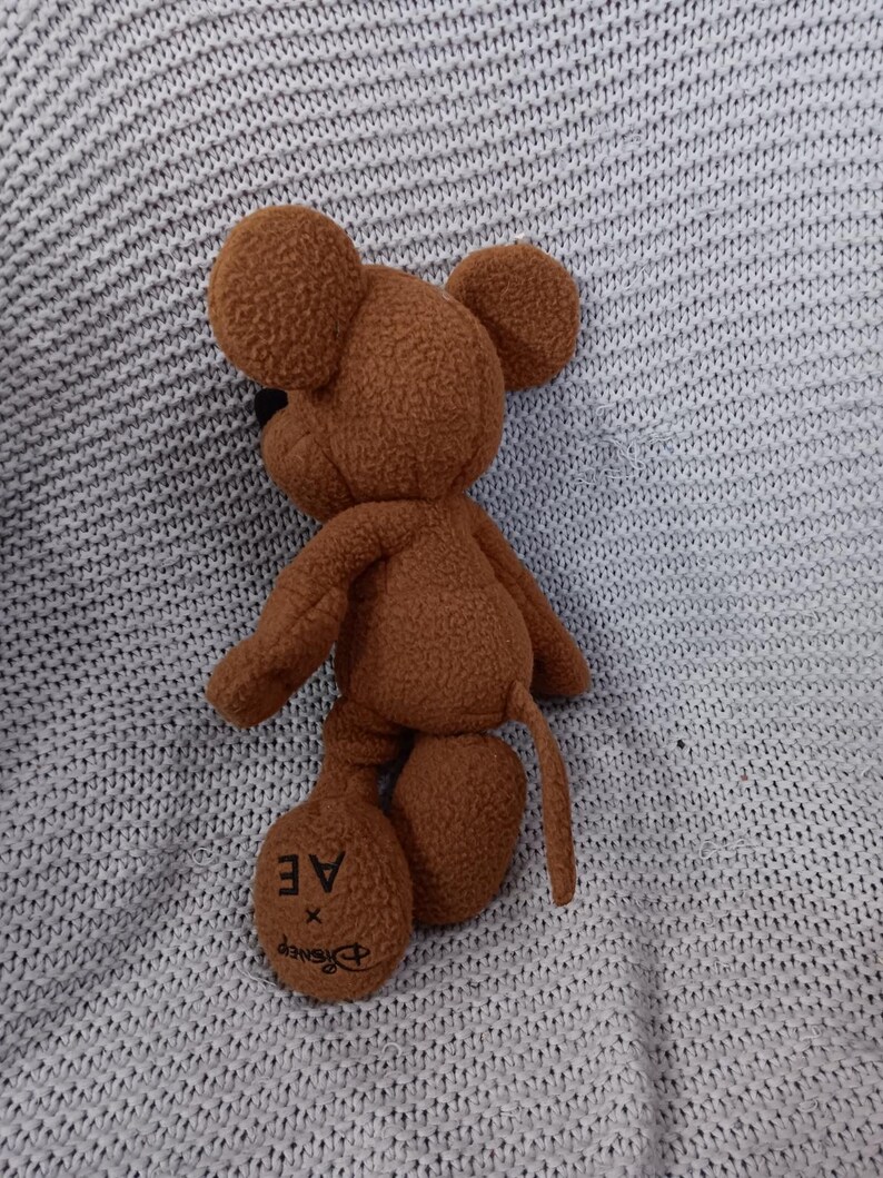 K&ouml;nnte beinhalten: Ein braunes Mickey Mouse-Pl&uuml;schtier mit gro&szlig;en, runden Ohren, einer schwarzen Nase und einem langen Schwanz. Auf einem Fu&szlig; des Spielzeugs steht "AE x Disney". Das Spielzeug liegt auf einer hellgrauen Strickoberfl&auml;che.